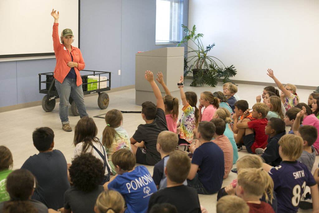 Woman in an orange sweater raises her hand in front of a group of kids seated on the floor.