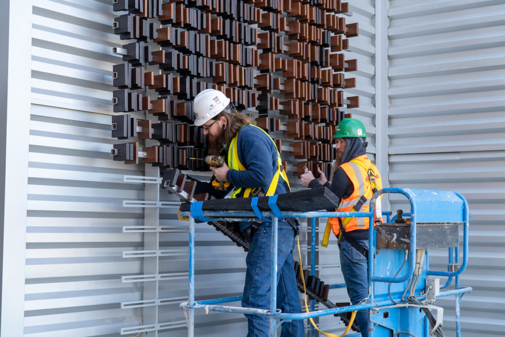 Two men in construction gear on a lift attach wood blocks to a wall.