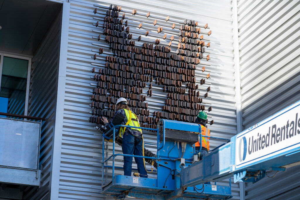 Two men on a lift work below a half finished horse head made of wood blocks.
