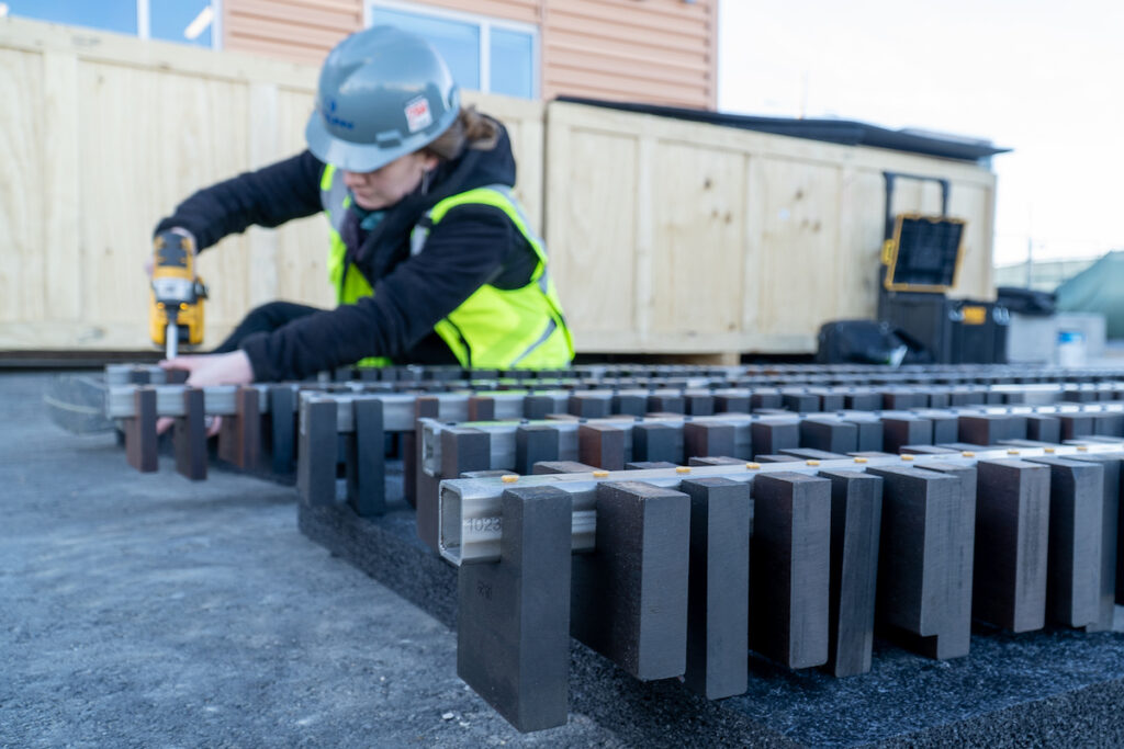 A woman in construction gear drills wood blocks onto a metal bar.