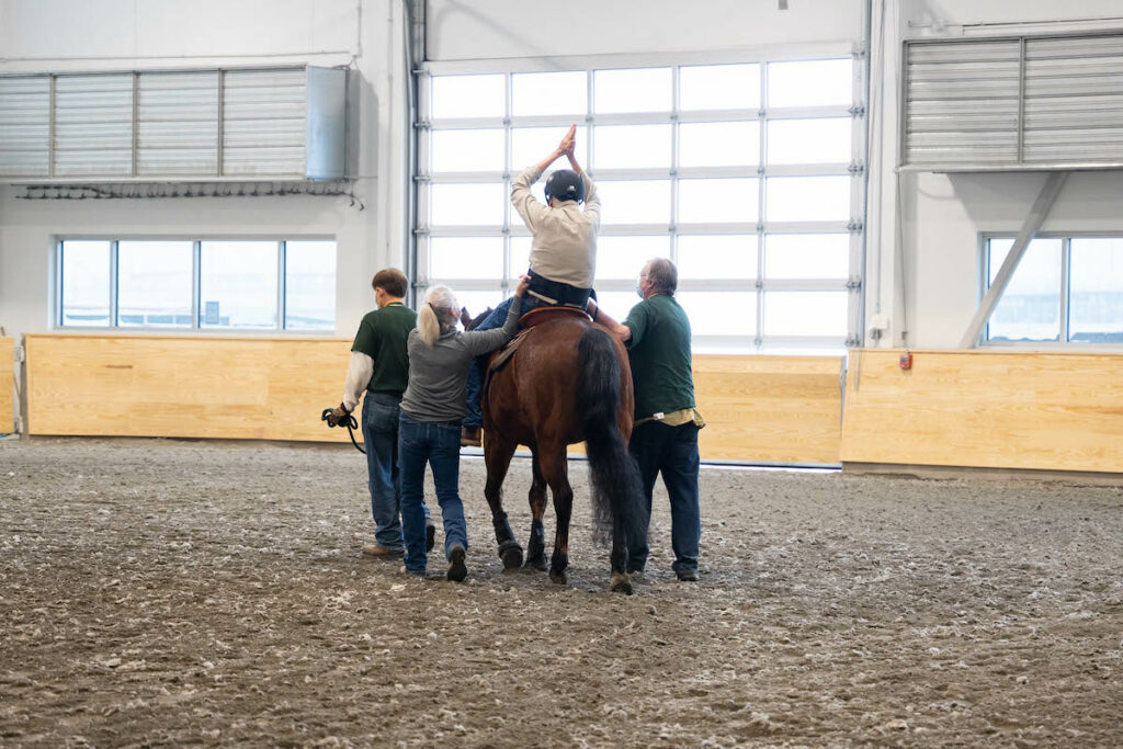 A man sits on a brown horse with his hands in the air and three people standing on the ground.