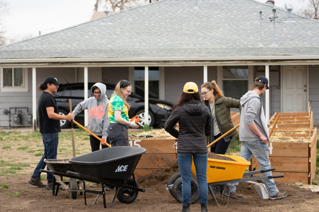 Five people stand around raised garden beds, wheelbarrows, and piles of mulch.