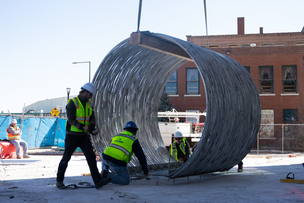 Construction workers attach a large metal sculpture shaped like a hay bale to a cement pad.