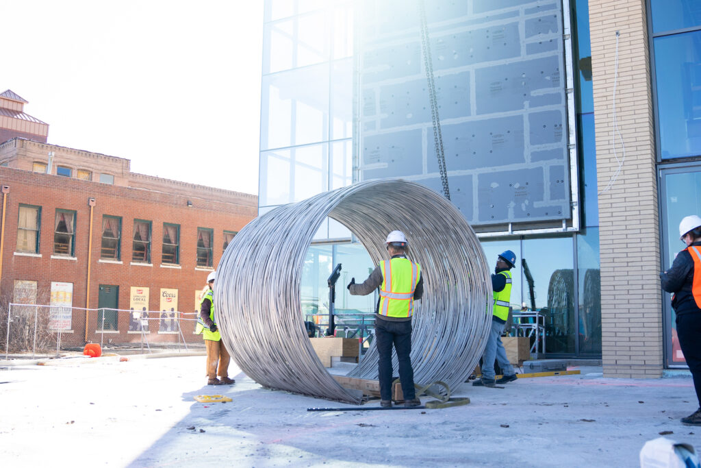 Construction workers install a large metal sculpture shaped like a hay bale