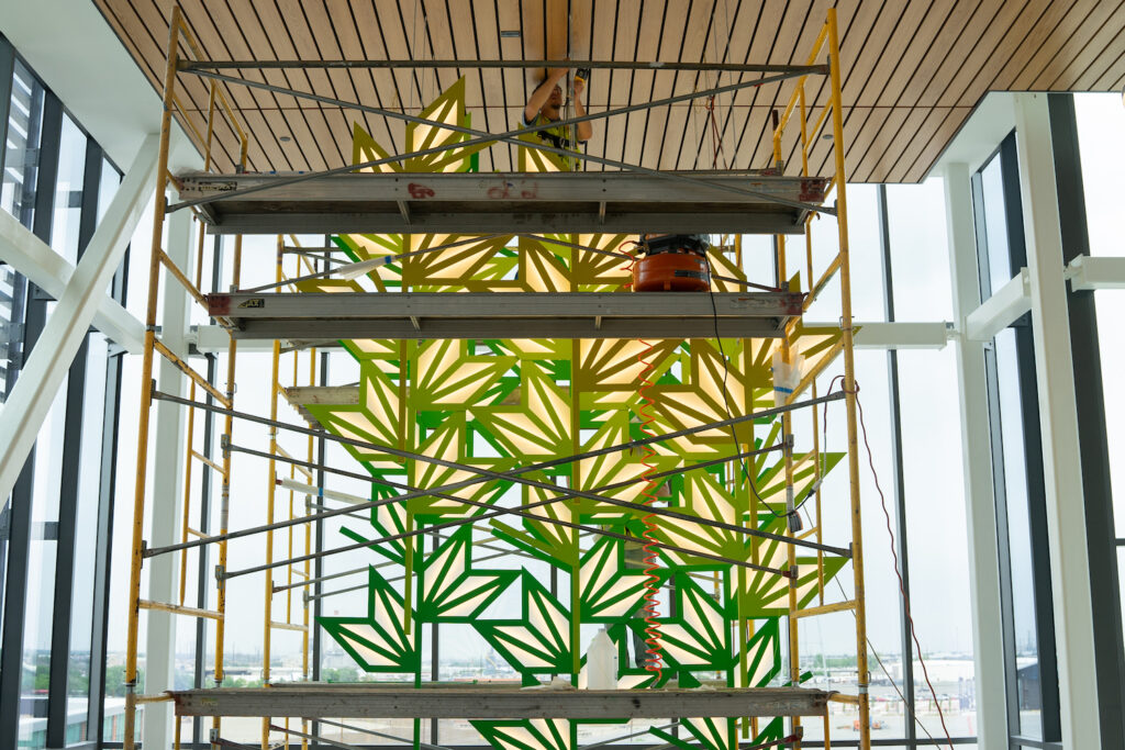 A person on a scaffold installs an art piece of green and yellow geometric leaf shapes.