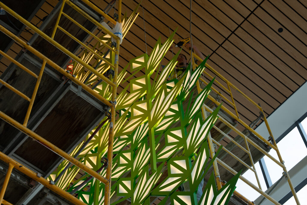 A person on a scaffold installs an art piece of green and yellow geometric leaf shapes