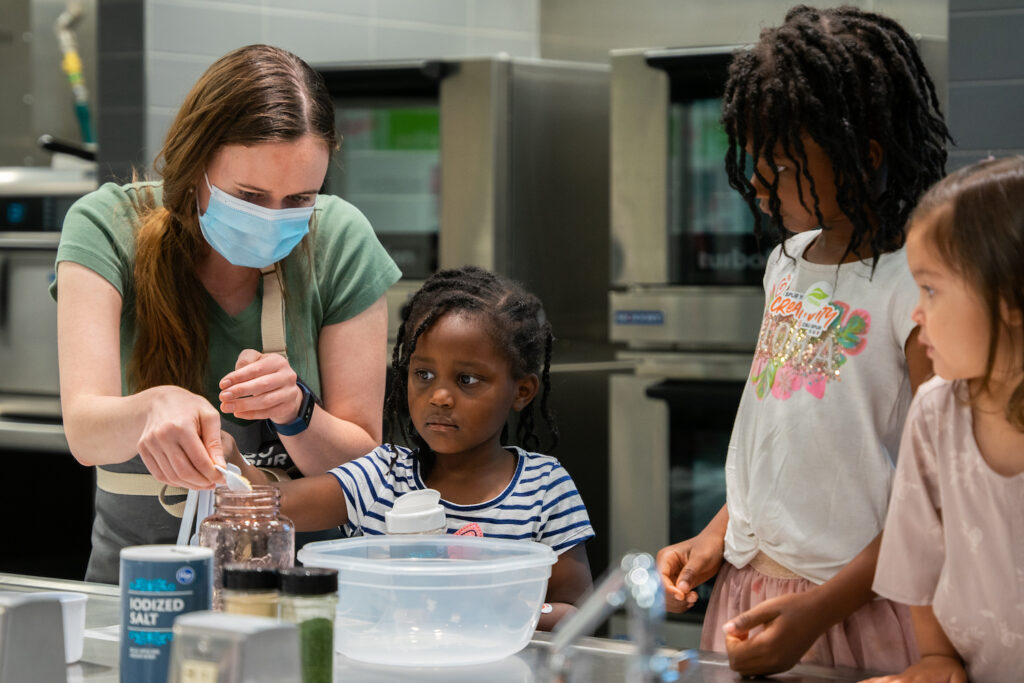 A woman helps three younger girls add ingredients to a jar.