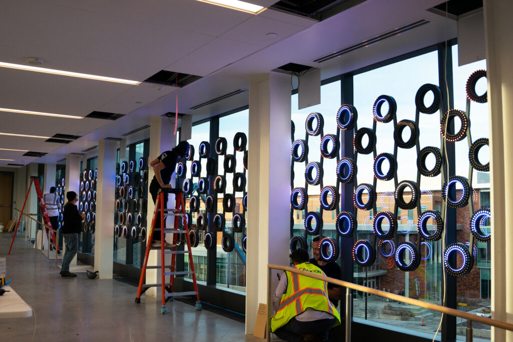 Crews in hard hats install an art installation on glass windows.
