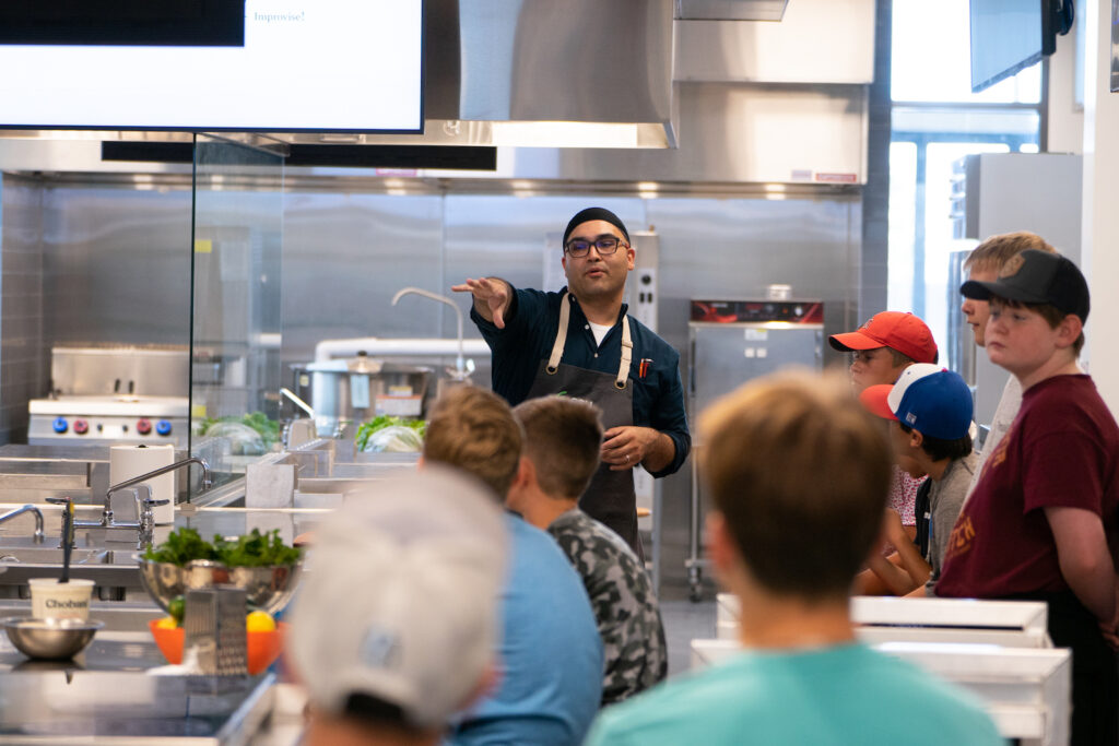 A man directs a cooking class in an industrial kitchen.
