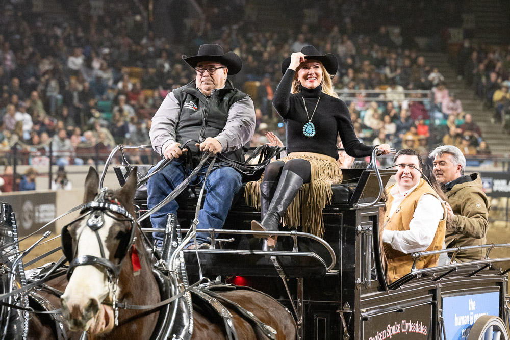 Two people, including CSU President Amy Parsons in western wear, ride on a stagecoach in a rodeo arena.