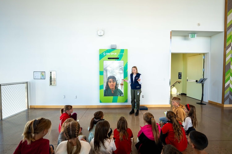 A woman in a Spur vest talks to a group of children seated on the floor in front of the wall-mounted CSU Spur Portal screen.