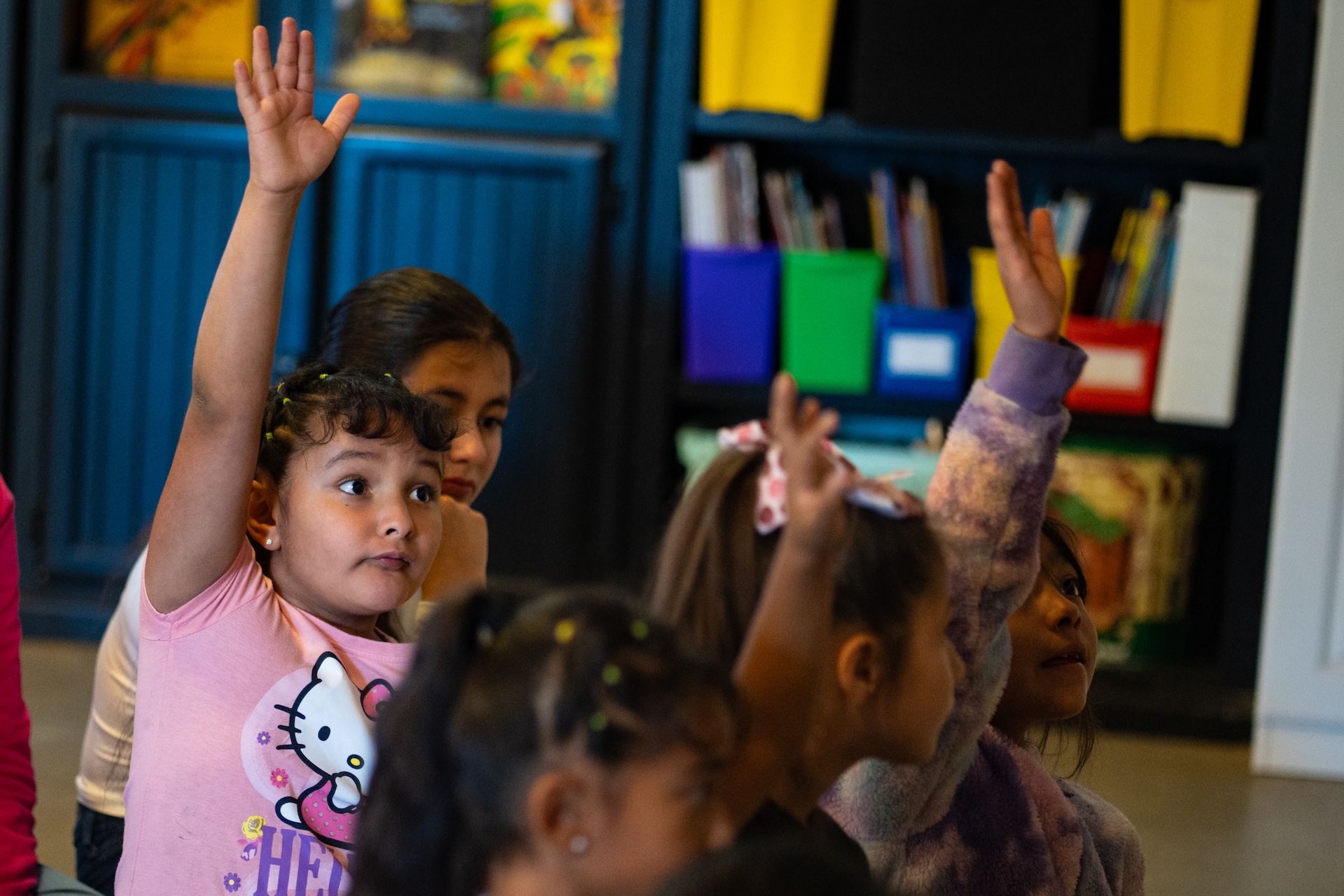 A girl in a pink Hello Kitty shirt raises her hand among a group of kids.