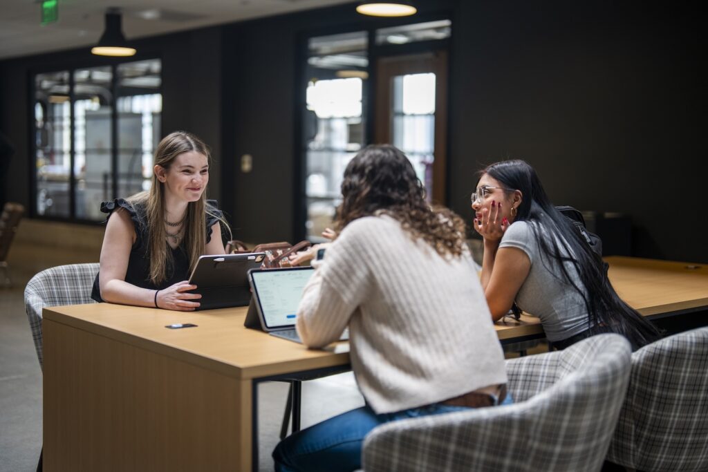 Three students work on laptops at a shared table.